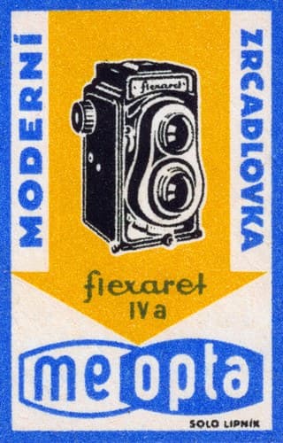 Černý dvouoký zrcadlový fotoaparát na žlutém poli. Kolem něj modré svislé nápisy, dole modrobílé logo Meopta.