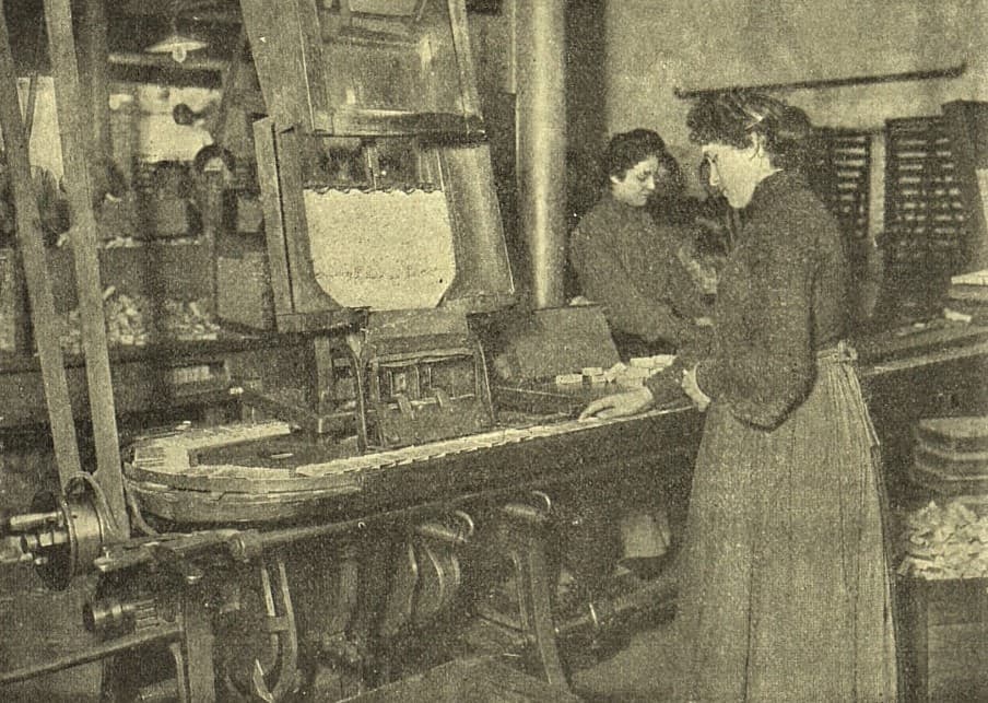 Dobová fotografie z roku 1907. Dvě dělnice v dlouhých sukních stojí u stroje na plnění zápalkových krabiček, na stole před nimi leží rozložené sirky.